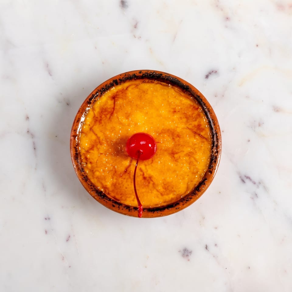 Overhead view of a creme brulee in a terracotta dish, garnished with a single red cherry.