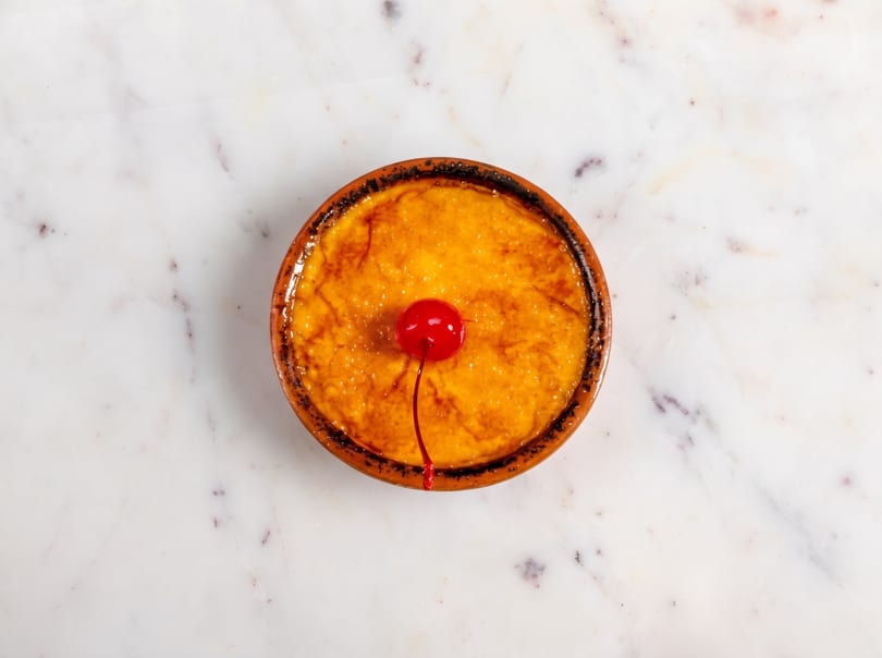 Overhead view of a creme brulee in a terracotta dish, garnished with a single red cherry.