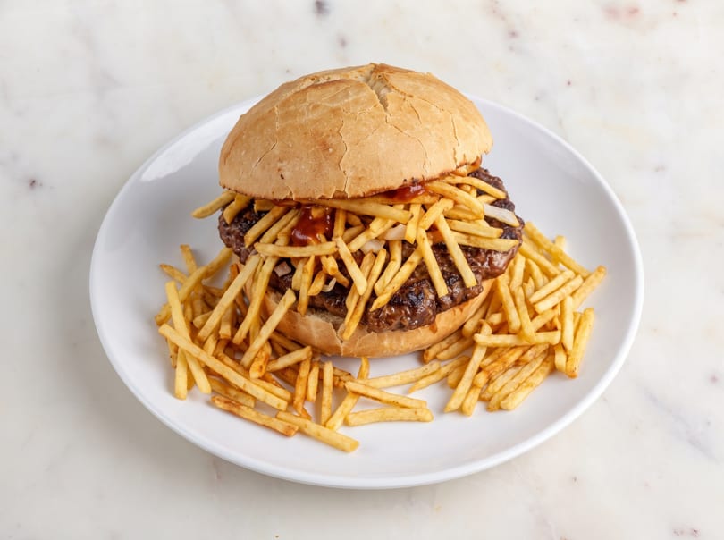 A large burger with fries and sauce on a white bun, surrounded by more fries on a white plate.